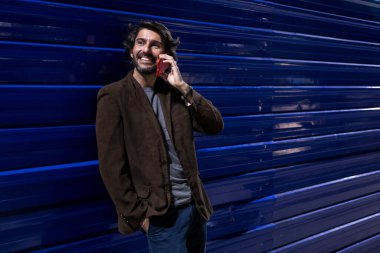 Gece vakti akıllı telefon kullanan genç bir adam şehir ışıklarının olduğu mavi bir duvara yaslanıyor. Gece vakti akıllı telefon kullanan genç adam, arka planda şehir manzarası olan mavi bir duvara yaslanıyor.. 