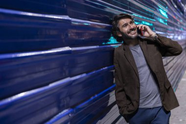 Gece vakti akıllı telefon kullanan genç bir adam şehir ışıklarının olduğu mavi bir duvara yaslanıyor. Gece vakti akıllı telefon kullanan genç adam, arka planda şehir manzarası olan mavi bir duvara yaslanıyor.. 