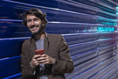 Gece vakti akıllı telefon kullanan genç bir adam şehir ışıklarının olduğu mavi bir duvara yaslanıyor. Gece vakti akıllı telefon kullanan genç adam, arka planda şehir manzarası olan mavi bir duvara yaslanıyor.. 