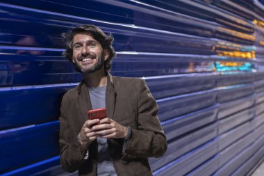 Gece vakti akıllı telefon kullanan genç bir adam şehir ışıklarının olduğu mavi bir duvara yaslanıyor. Gece vakti akıllı telefon kullanan genç adam, arka planda şehir manzarası olan mavi bir duvara yaslanıyor.. 