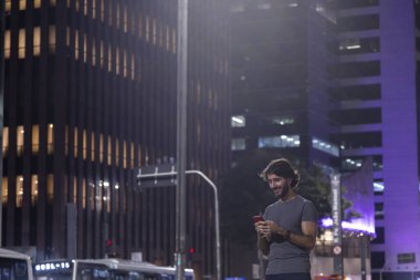 Gece vakti akıllı telefon kullanan genç bir adam. Şehir manzarası ve arka planda araba trafiği var.. 