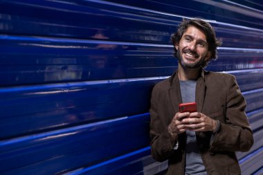 Gece vakti akıllı telefon kullanan genç bir adam şehir ışıklarının olduğu mavi bir duvara yaslanıyor. Gece vakti akıllı telefon kullanan genç adam, arka planda şehir manzarası olan mavi bir duvara yaslanıyor.. 