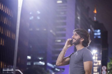 Gece vakti akıllı telefon kullanan genç bir adam. Şehir manzarası ve arka planda araba trafiği var.. 