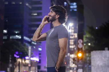 Gece vakti akıllı telefon kullanan genç bir adam. Şehir manzarası ve arka planda araba trafiği var.. 