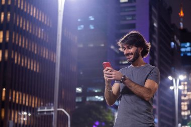 Gece vakti akıllı telefon kullanan genç bir adam. Şehir manzarası ve arka planda araba trafiği var.. 