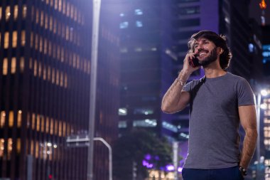 Gece vakti akıllı telefon kullanan genç bir adam. Şehir manzarası ve arka planda araba trafiği var.. 