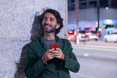 Gece vakti akıllı telefon kullanan genç bir adam. Şehir manzarası ve arka planda araba trafiği var.. 