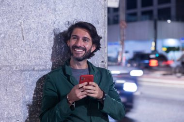 Gece vakti akıllı telefon kullanan genç bir adam. Şehir manzarası ve arka planda araba trafiği var.. 
