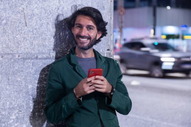 Gece vakti akıllı telefon kullanan genç bir adam. Şehir manzarası ve arka planda araba trafiği var.. 