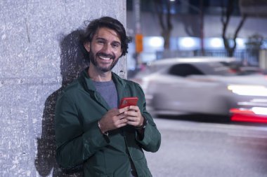 Gece vakti akıllı telefon kullanan genç bir adam. Şehir manzarası ve arka planda araba trafiği var.. 