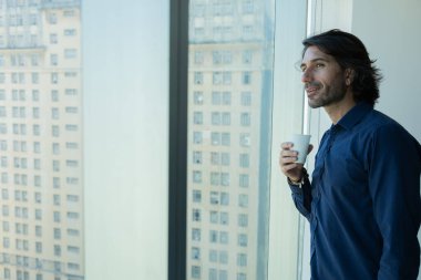 Ofiste elinde bir fincan kahve tutan ve içen Latin Amerikalı genç bir adamın portresi. Arka planda şehir manzarası olan dışarı bakıyor..  