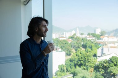 Ofiste elinde bir fincan kahve tutan ve içen Latin Amerikalı genç bir adamın portresi. Arka planda şehir manzarası olan dışarı bakıyor..  