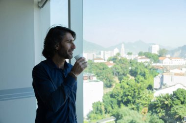 Ofiste elinde bir fincan kahve tutan ve içen Latin Amerikalı genç bir adamın portresi. Arka planda şehir manzarası olan dışarı bakıyor..  