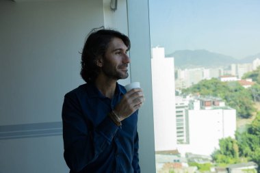 Ofiste elinde bir fincan kahve tutan ve içen Latin Amerikalı genç bir adamın portresi. Arka planda şehir manzarası olan dışarı bakıyor..  