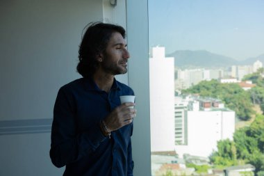 Ofiste elinde bir fincan kahve tutan ve içen Latin Amerikalı genç bir adamın portresi. Arka planda şehir manzarası olan dışarı bakıyor..  