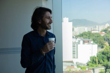 Ofiste elinde bir fincan kahve tutan ve içen Latin Amerikalı genç bir adamın portresi. Arka planda şehir manzarası olan dışarı bakıyor..  