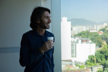 Ofiste elinde bir fincan kahve tutan ve içen Latin Amerikalı genç bir adamın portresi. Arka planda şehir manzarası olan dışarı bakıyor..  