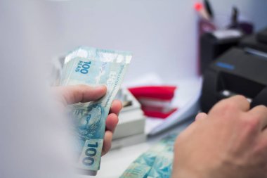 Değişim bürosu / bankada çalışan iş adamının insan eli. O para sayıyor ve bir müşteriye nakit para veriyor. Brezilya Reais 100 reais.
