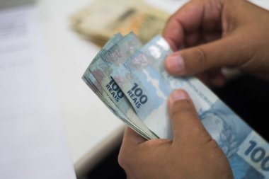 Değişim bürosu / bankada çalışan iş adamının insan eli. O para sayıyor ve bir müşteriye nakit para veriyor. Brezilya Reais 100 reais.