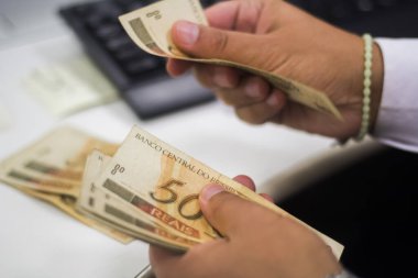 Değişim bürosu / bankada çalışan iş adamının insan eli. O para sayıyor ve bir müşteriye nakit para veriyor. Brezilyalı 50 reais fatura.