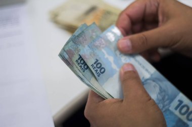 Değişim bürosu / bankada çalışan iş adamının insan eli. O para sayıyor ve bir müşteriye nakit para veriyor. Brezilya Reais 100 reais.