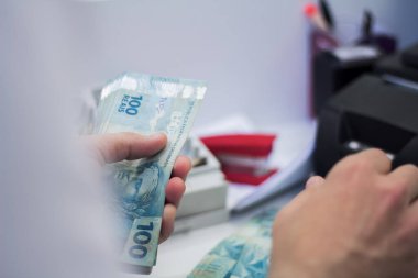Değişim bürosu / bankada çalışan iş adamının insan eli. O para sayıyor ve bir müşteriye nakit para veriyor. Brezilya Reais 100 reais.