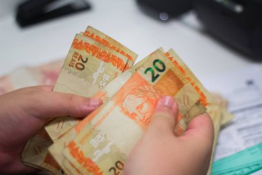 Değişim bürosu / bankada çalışan iş adamının insan eli. O para sayıyor ve bir müşteriye nakit para veriyor. Brezilya Reais - 20 reais fatura.