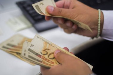 Değişim bürosu / bankada çalışan iş adamının insan eli. O para sayıyor ve bir müşteriye nakit para veriyor. Brezilyalı 50 reais fatura.