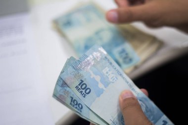Değişim bürosu / bankada çalışan iş adamının insan eli. O para sayıyor ve bir müşteriye nakit para veriyor. Brezilya Reais 100 reais.