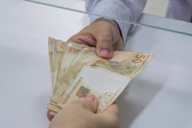 Değişim bürosu / bankada çalışan iş adamının insan eli. O para sayıyor ve bir müşteriye nakit para veriyor. Brezilyalı 50 reais fatura.