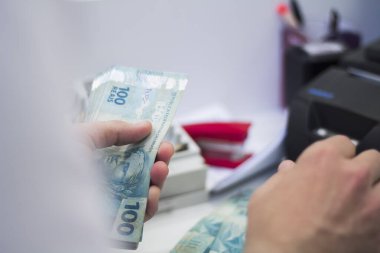 Değişim bürosu / bankada çalışan iş adamının insan eli. O para sayıyor ve bir müşteriye nakit para veriyor. Brezilya Reais 100 reais.