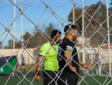 Hakem ve oyuncular güneşli bir günde sentetik çim sahasında futbol oynuyorlar.