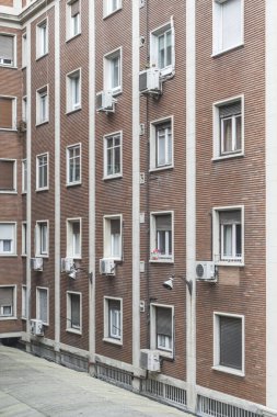 Açıkta kil renkli tuğla, klimalı pencereli ve beyaz taşlı bir binanın ön cephesi.