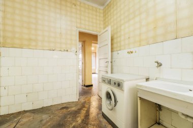 Cocina vieja, sucia y destartalada con suelo oscuro, azulejos blancos y papel amarillo en las paredes