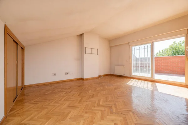 Empty living room with fitted wardrobes, parquet floors, sloping ceilings and access to a terrace with a fence