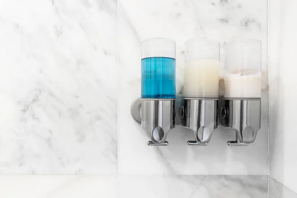 Soap, shampoo and softener dispensers on the wall of a shower tiled in white marble