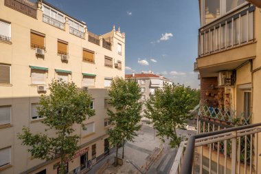 Genç ağaçlarla dolu bir cadde, bitkilerle dolu balkonlar ve bulutlarla dolu bir gökyüzü.