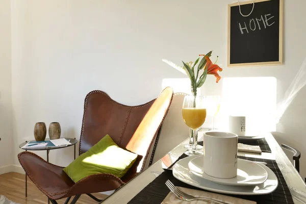 A corner of a small living room with a table with orange juice, some flowers and a leather armchair in a vacation rental apartment