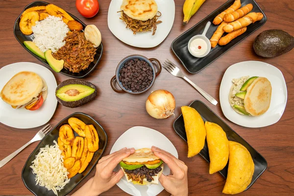 A table full of plates of Latin and Venezuelan food with hands holding an arepa