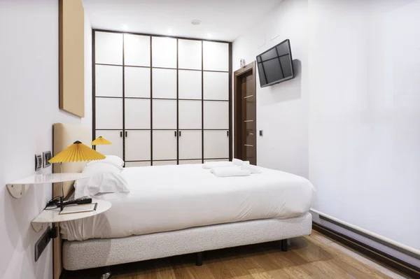 Bedroom with a double bed with a white feather duvet with white shelves as bedside tables with twin lamps and a large built-in wardrobe with white wooden doors with bars and oak parquet floors