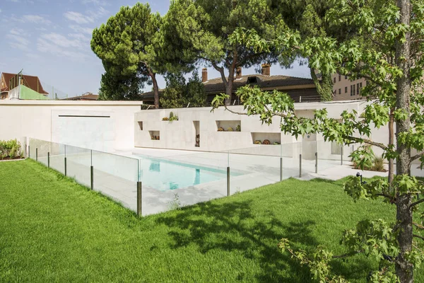 Beautiful swimming pool of a luxury detached house with marble tiles and a wall with niches to accommodate outdoor plants, glass fence with metal stilts on the lawn, palm trees and plants