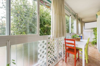 Closed terrace of a house with a white table and indoor hanging plants, white wooden latticework and views of a wooded garden
