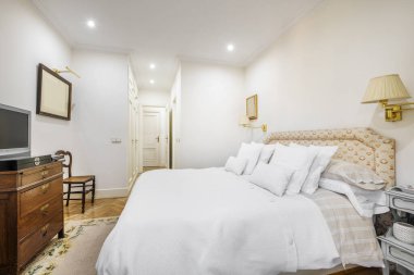 A room with a double bed decorated in a classic style with a headboard upholstered in fabrics and many cushions of different sizes with white covers