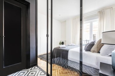 A room with a double bed with many cushions and a balcony overlooking the street seen through the glass of a screen