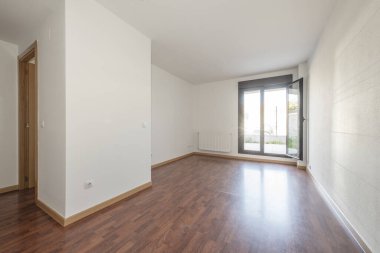 Empty room with dark reddish wooden floor, smooth white walls, and access to a terrace with a dark anodized aluminum door