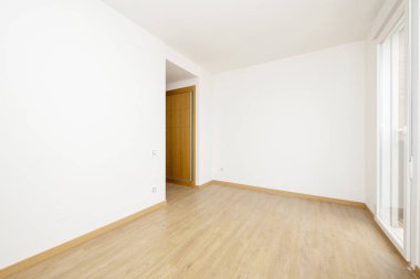Empty room with light wooden floors, white aluminum radiators, smooth white painted walls, a built-in wardrobe in the access corridor and a white aluminum and glass balcony door
