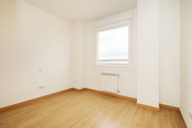Empty room with light oak wooden floors, white aluminum radiators, white painted plain walls and a single window with a roller blind