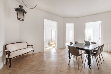 Living room with an elongated armchair in a corner, a vintage wooden table with wheels on its legs and several accesses to other rooms without doors