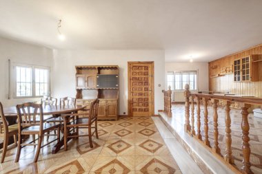 Living room with double-height stoneware floors, a wooden railing, dining table with matching chairs, pine paneled service door and wall with a tongue-and-groove wood mural