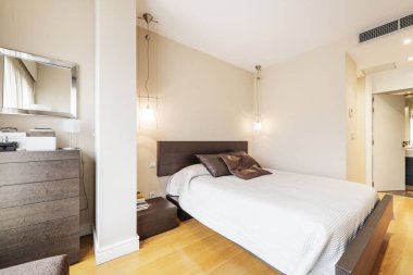 Bedroom with a double bed with a brown leather upholstered headboard, matching cushions, white bedspread, square mirror hanging on the wall over a chest of drawers